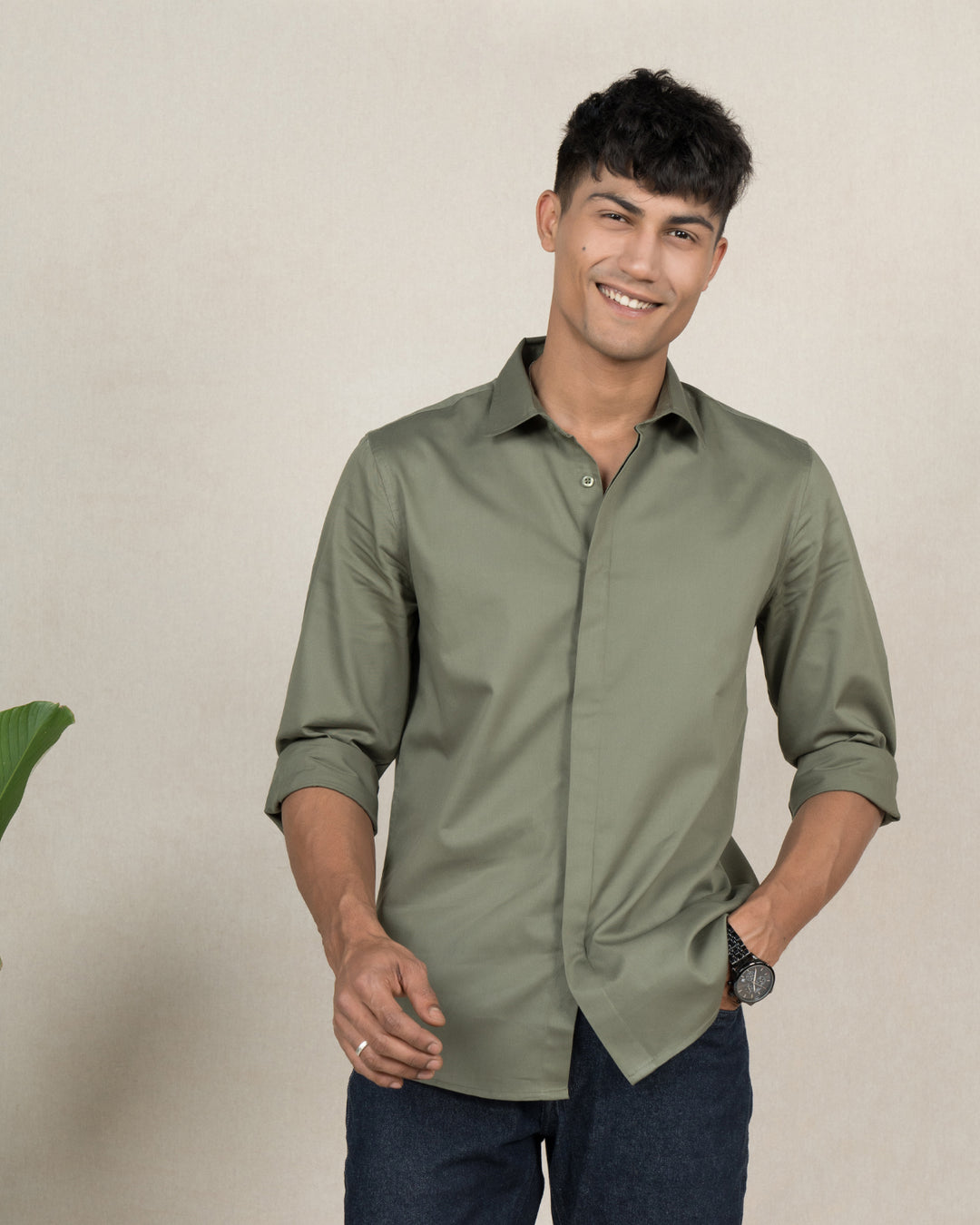 A young person with short dark hair is smiling, wearing a timeless Dark Sage - Classic Shirt made from premium cotton that offers luxury softness. Their sleeves are rolled up, and they pair it with dark jeans. With their right hand in their pocket, they stand against a neutral background with a small green plant to the left.