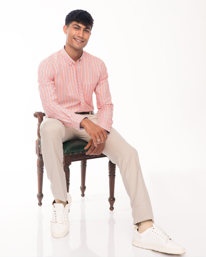 A man sits on a wooden chair with a green seat against a white background. He is wearing a Quartz - Oxford Shirt made of premium cotton, beige pants, and white sneakers. He is smiling, with one hand resting on his thigh and the other on the chair, embodying eco-conscious designs in his attire.