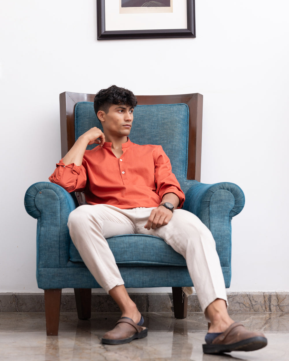 A young man with curly hair is standing indoors, wearing a Scarlatto - Short Kurta and beige pants. He is looking to his right with his left hand in his pocket. Behind him are a teal armchair, a framed picture on the wall, and a bookshelf with several books.