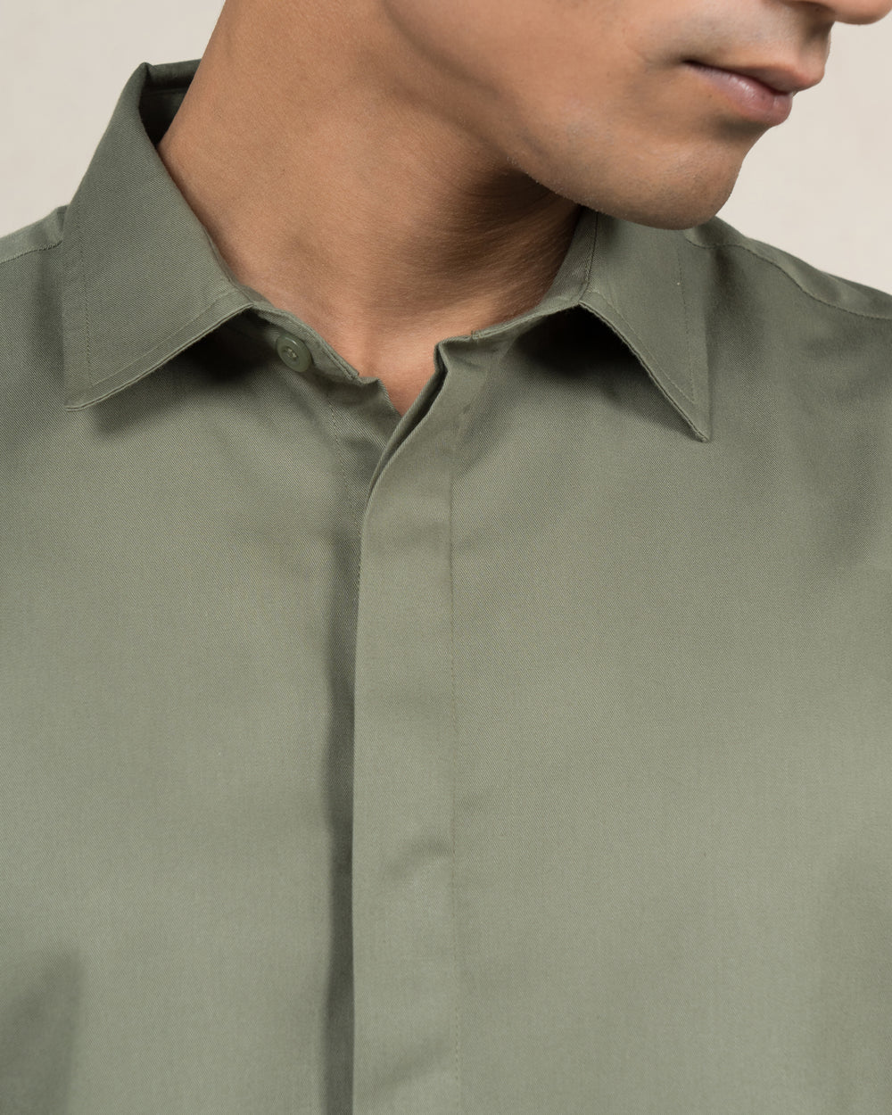 A young person with short dark hair is smiling, wearing a timeless Dark Sage - Classic Shirt made from premium cotton that offers luxury softness. Their sleeves are rolled up, and they pair it with dark jeans. With their right hand in their pocket, they stand against a neutral background with a small green plant to the left.