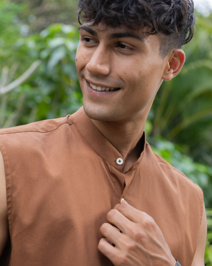 A man with short, curly hair wearing sunglasses, a sleeveless brown button-up Brunn - Sleeveless Shirt, and beige pants stands outdoors. He is looking down with his hands in his pockets. A backdrop of lush green foliage and trees is visible, showcasing his effortless style.
