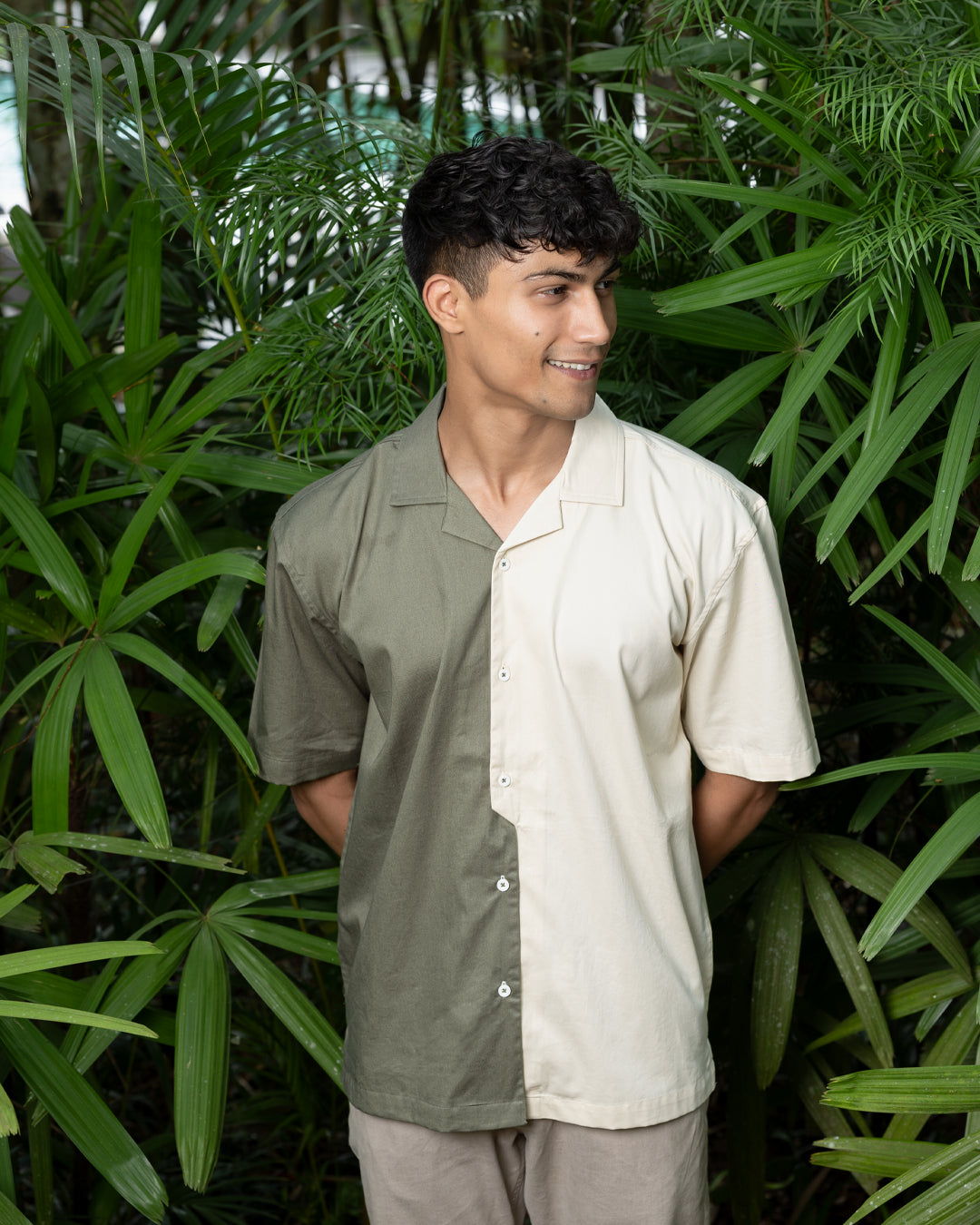 A man with curly dark hair smiles while standing in front of greenery. He is wearing a short-sleeved Dark Sage Moitie - Cuban Collar Shirt that is olive green on one side and cream on the other, paired with light-colored pants. He stands with one hand in his pocket.